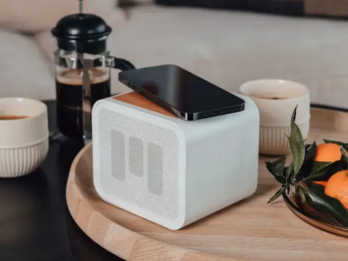 En smartphone hviler på en hvid trådløs højttaler på en træbakke ved siden af kaffekopper, en stempelkande og en lille tallerken med mandariner med blade.