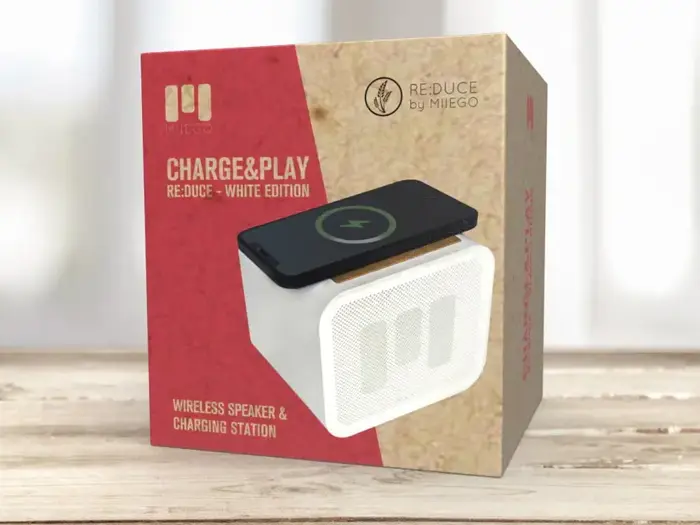 En produktkasse på en træoverflade, der viser den trådløse MIEGO CHARGE&PLAY-højttaler og opladningsstation, med en telefon, der oplades på toppen, og etiketten RE:DUCE - WHITE EDITION.