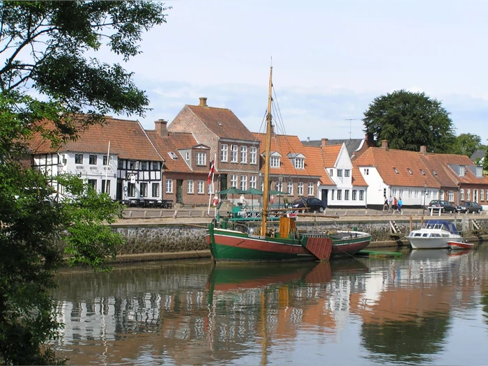En grøn og rød træbåd, der ligger fortøjet i en kanal foran en række traditionelle europæiske huse med orange tegltage.