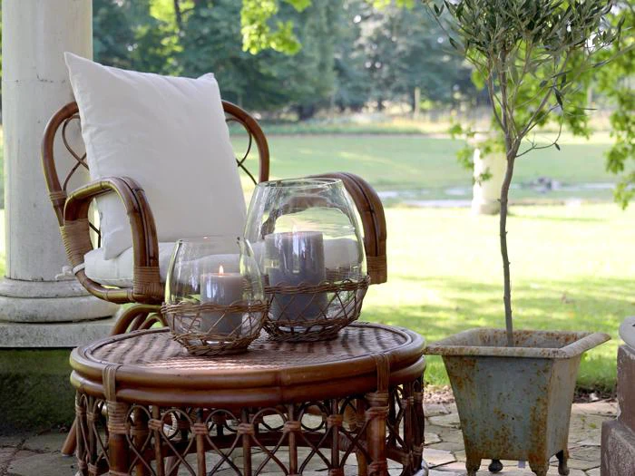 En kurvestol med en hvid hynde står ved siden af et rundt kurvebord med stearinlys i glasholdere på en terrasse med en potteplante og en grøn græsplæne i baggrunden.