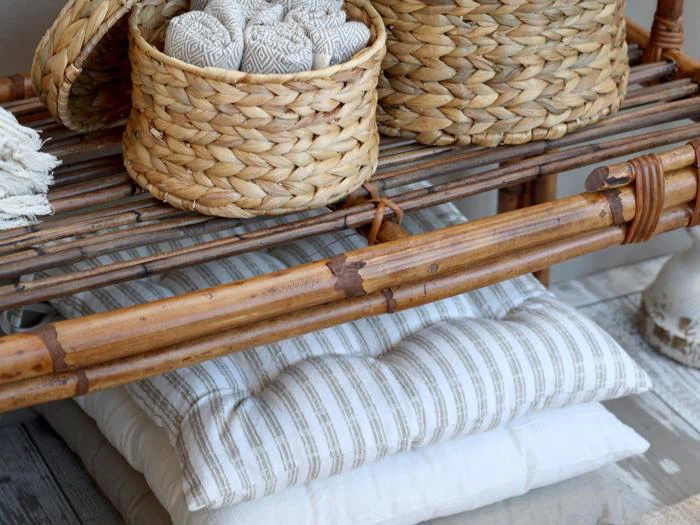 Flettede kurve med mønstrede kugler står på en bambushylde over stablede hvide og stribede puder på et lyst trægulv.