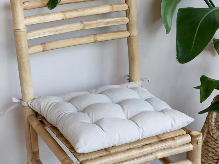 En lys, tuftet pude placeret på en bambusstol med grønne blade delvist synlige på højre side.