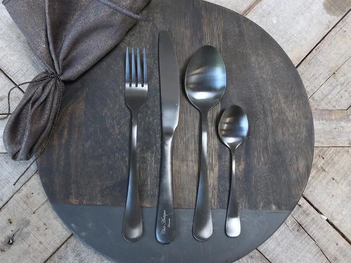 Et sæt matsort bestik - gaffel, kniv, spiseske og teske - ligger pænt på et rundt, mørkt træbræt med en foldet serviet ved siden af.
