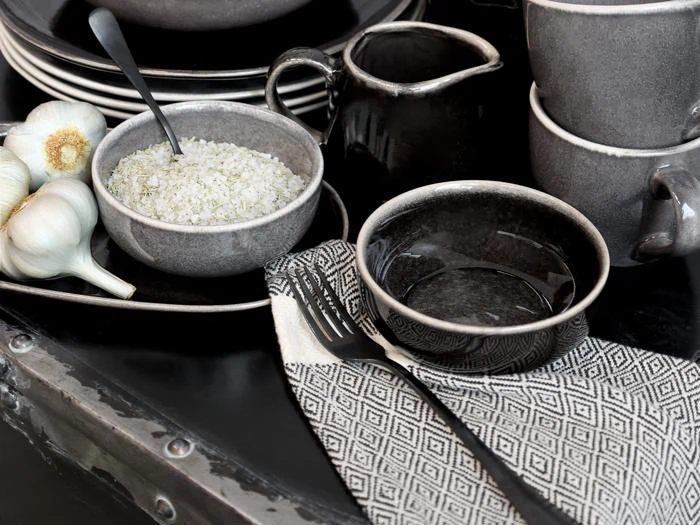 Et dækket bord med grå keramikfade, en skål med groft salt med en ske, en mønstret serviet med bestik, hele hvidløgsløg og stablede kopper på en mørk overflade.