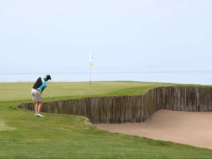 En golfspiller slår et slag fra græsset nær en bunker mod en green med et flag med udsigt til et roligt vandområde i baggrunden.