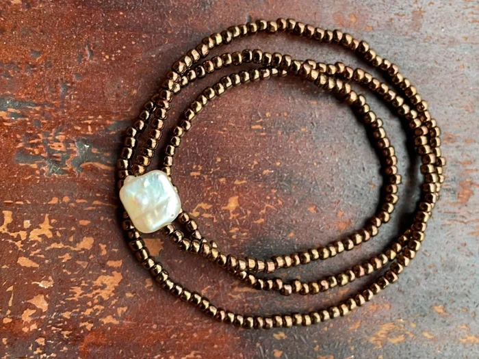 En perlekæde med små bronzeperler og en enkelt stor, uregelmæssig hvid perle på en rustik, brun overflade.