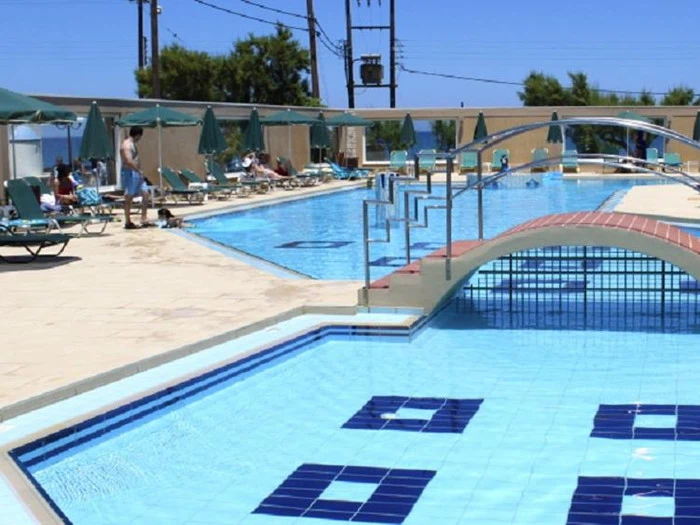 Udendørs swimmingpoolområde med en lille gangbro, liggestole, grønne parasoller og folk, der slapper af ved poolen under en klar blå himmel.