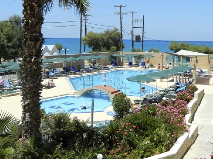 Solrigt poolområde med liggestole, parasoller og en lille bro over poolen med udsigt til havet og blomster i forgrunden.