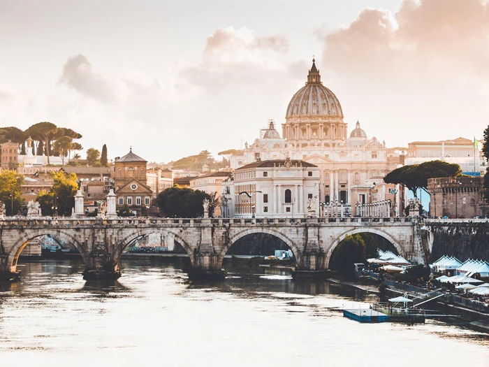 En naturskøn udsigt over Peterskirken i Vatikanstaten med Tiberen og en buet stenbro i forgrunden under en delvis overskyet himmel.