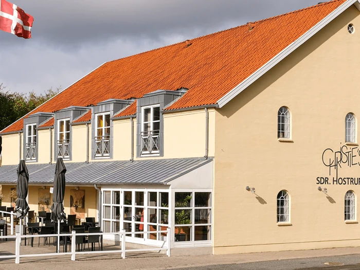 En gul, toetagers bygning med rødt tegltag, udeservering og store vinduer; et dansk flag er synligt i øverste venstre hjørne.