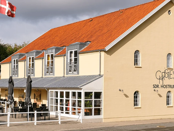 En gul toetagers bygning med rødt tegltag, udeservering, store vinduer og et delvist synligt flag under en overskyet himmel.