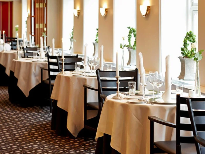 Elegant restaurant med pænt dækkede borde, hvide duge, sorte stole, stearinlys, glas og potteplanter ved store vinduer, der lukker naturligt lys ind.