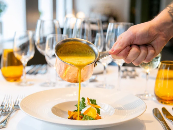 En hånd hælder gul sauce fra en lille gryde over et fad med grøntsager, der er dækket på et elegant bord med glas og sølvtøj.