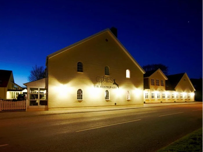 En varmt oplyst, lysegul bygning med navnet Christies Sdr. Hostrup Kro står langs en tom vej i skumringen under en dybblå himmel.