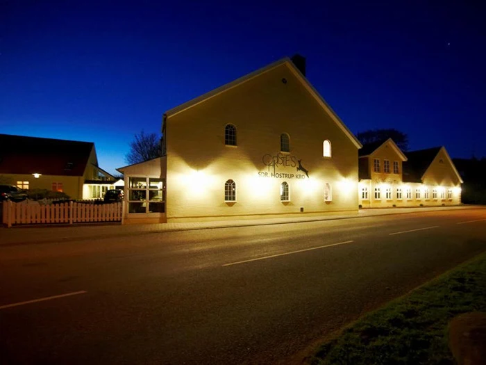 En veloplyst bygning med skiltet Op Tersk For Hosterup Kro står ved en stille vej i skumringen med en mørkeblå himmel og et hvidt stakit.