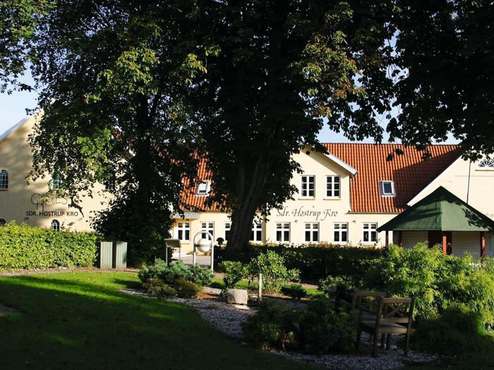 En gul kro med rødt tegltag står bag træer og en grøn have med bænke, stier og buske på en solrig dag.