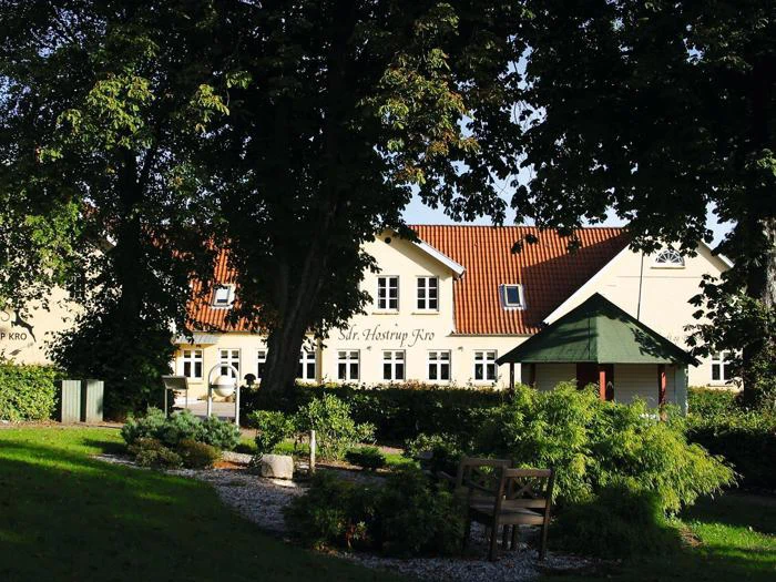 En cremefarvet bygning med rødt tegltag ligger bag træer og grønne områder med en havebænk og et lille lysthus i forgrunden.