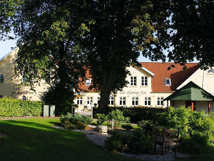 En solbrun bygning med røde tagsten, omgivet af grønne træer og buske, med bænke og en grussti i en solbeskinnet have.