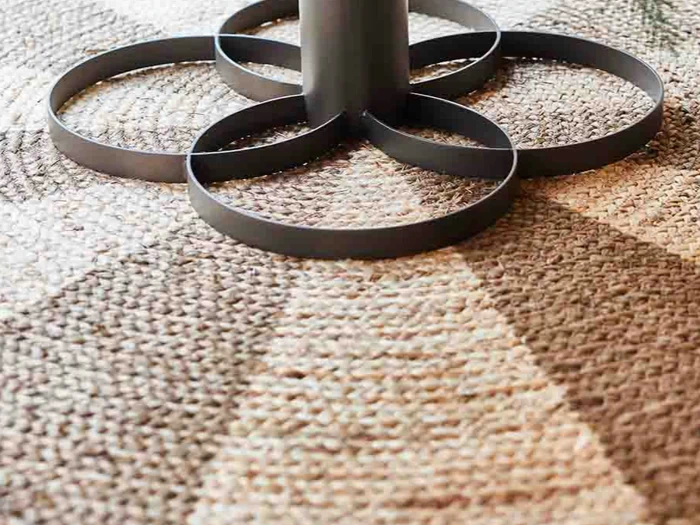 Nærbillede af en metalbordfod med fire cirkulære støtter på et struktureret, mønstret vævet tæppe i brune og beige nuancer.