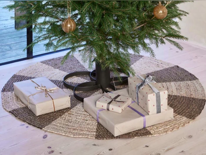 Et juletræ står på et vævet tæppe med flere indpakkede gaver i neutralt papir og bånd placeret nedenunder.