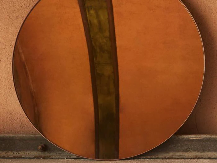 Et rundt spejl læner sig op ad en struktureret ferskenfarvet væg og reflekterer en lodret grønbrun stribe og en del af væggen.