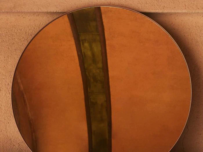 Et rundt spejl reflekterer en orangebrun væg og en lodret grønlig stribe, hvilket skaber en abstrakt komposition.