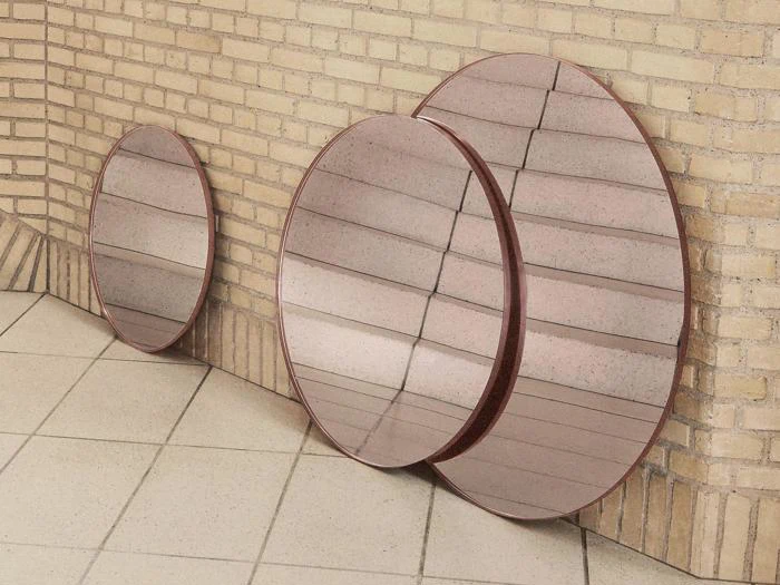 Tre runde spejle med rødlige rammer står op ad en lys murstensvæg på et flisegulv og reflekterer loftet og vægfliserne.