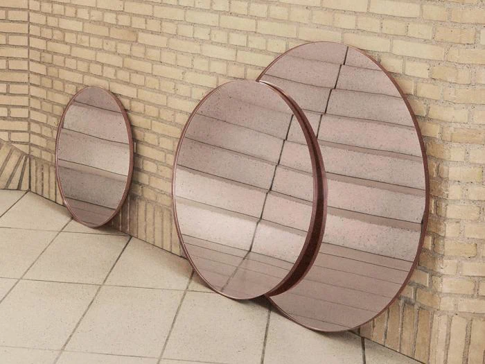 Tre runde spejle med rødlige rammer læner sig op ad en solbrun murstensvæg på et flisegulv og reflekterer væg- og gulvfliserne.