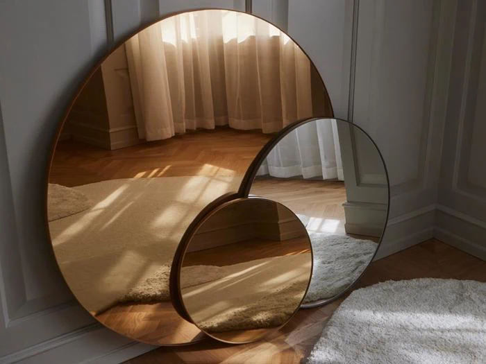Tre runde spejle i forskellige størrelser med kobberfarvede rammer læner sig op ad en hvid væg og reflekterer sollys, gardiner og en del af et tæppe på et trægulv.