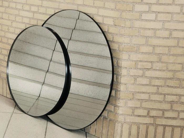 To runde spejle med sorte rammer står lænet op ad en lys murstensvæg og reflekterer klinkegulvet og murstensmønsteret.