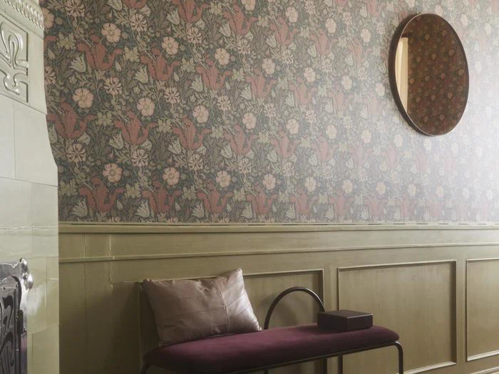 Et rum med blomstret tapet, grønne paneler, et rundt vægspejl og en bænk med en beige pude og en mørk kasse på.
