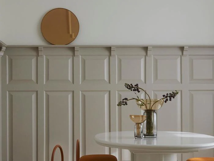 En minimalistisk spiseplads med beigefarvede vægge, et rundt spejl, et hvidt bord, brune stole og glasvaser med blomster.