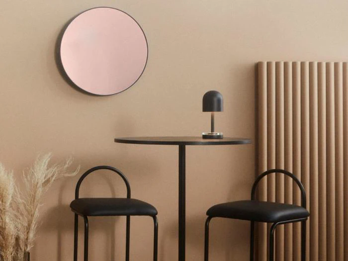 Minimalistisk rum med et lille rundt bord, to sorte stole, et rundt rosa spejl, en lille lampe, en ribbet radiator og dekorativt tørret græs mod beige vægge.