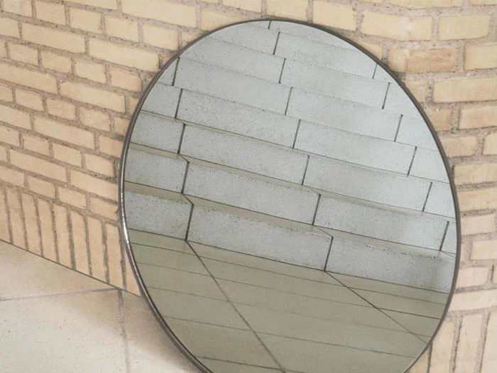 Et rundt spejl læner sig op ad en murstensvæg og reflekterer en flisebelagt trappe med lyse trin og søjler.