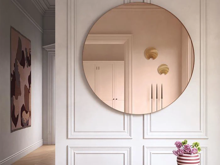 Et stort rundt spejl på en væg med hvide paneler afspejler en gang med skabe og to muslingeformede lampetter; en vase med lyserøde blomster står under spejlet.