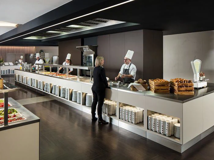 Et moderne cafeteria med kokke i hvide uniformer, der serverer mad bag en disk, mens en kvinde i forretningstøj taler med en af dem. Tallerkener og kopper er pænt stablet under disken.