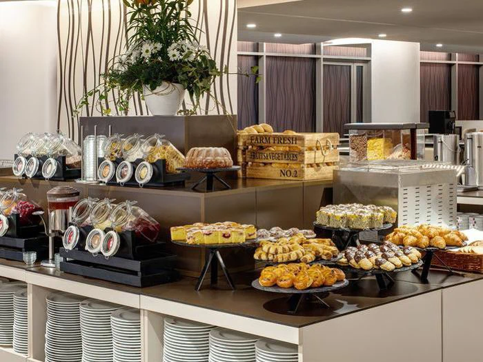 På et buffetbord står forskellige kager, croissanter, glas med syltetøj og stablede tallerkener med et stort blomsterarrangement i baggrunden.