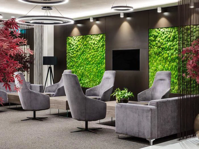 Moderne lounge med grå lænestole, træborde, røde planter, grønne vægpaneler med mos, ringkroner og et tv monteret på en mørk væg.