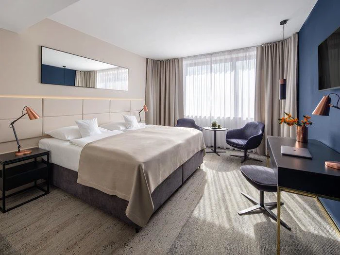 Moderne hotelværelse med en stor dobbeltseng, to stole, et rundt bord ved vinduet, et skrivebord med en lampe og neutral indretning. Naturligt lys strømmer ind gennem gennemsigtige gardiner.