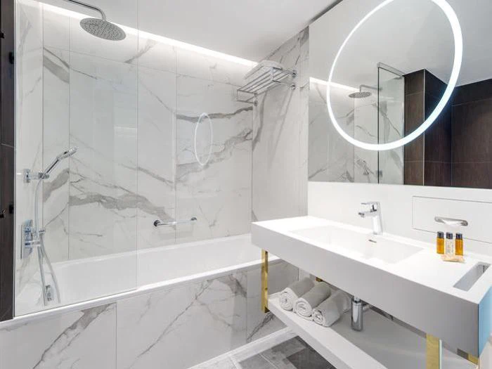 Moderne badeværelse med hvide marmorvægge, badekar med bruseafskærmning i glas, dobbelt håndvask og et rundt, oplyst spejl over håndvasken. Håndklæder og toiletartikler er pænt arrangeret.