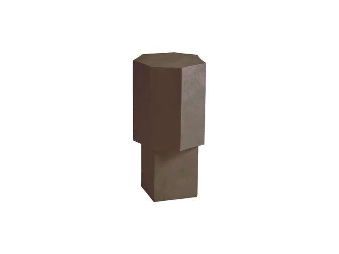 En høj, mørk, geometrisk skulptur med en ottekantet top og en rektangulær base, placeret foran en ensfarvet hvid baggrund.