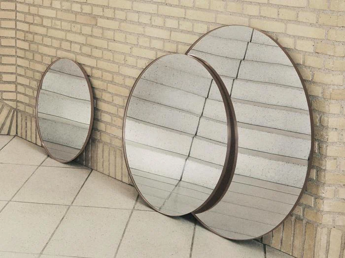 Tre runde spejle i forskellige størrelser står lænet op ad en lys murstensvæg på et flisegulv og reflekterer væg- og gulvmønsteret.