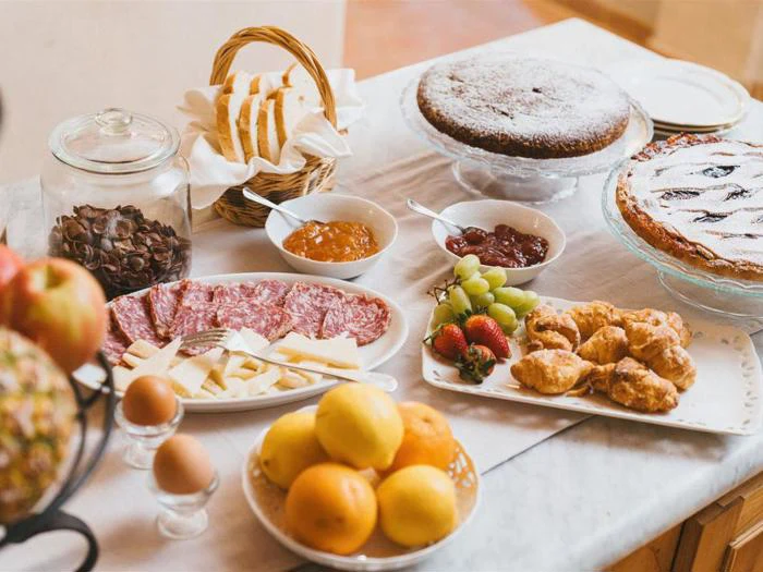 Et morgenbord med skiveskåret brød, chokoladekage, frugttærte, kødpålæg, ost, croissanter, frugt, kogte æg, syltetøj og et glas chokolademüsli.