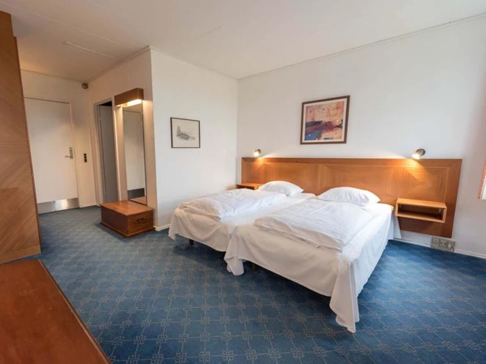 Et hotelværelse med to enkeltsenge skubbet sammen, hvidt sengetøj, blåt gulvtæppe, træmøbler, kunstværker på væggen og en dør ud til gangen.