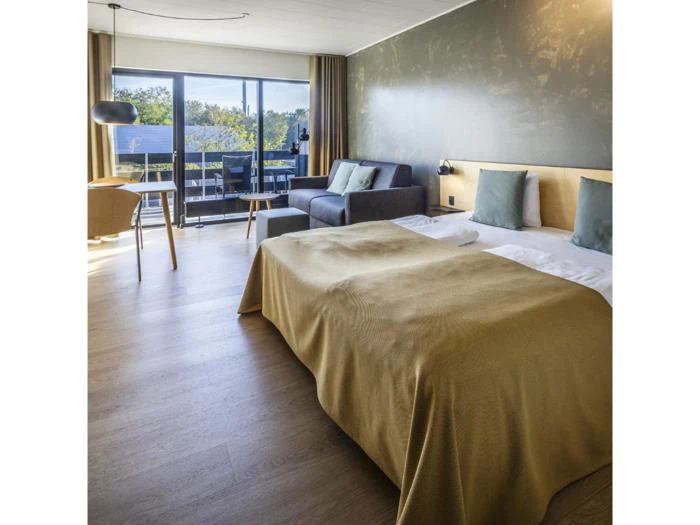 Moderne hotelværelse med dobbeltseng, sofa, lille bord med stole, stort vindue og balkon med grønt udenfor. Værelset har trægulv og dæmpede grønne og gule toner.