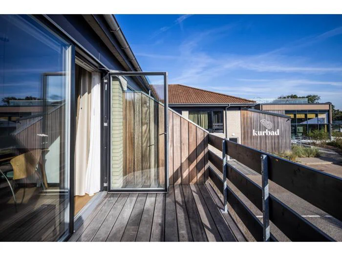 En træbalkon med en åben glasdør med udsigt til moderne bygninger og et skilt, hvor der står kurbad, under en klar blå himmel.