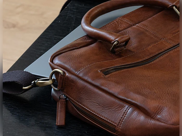 En brun lædertaske med lynlås hviler oven på en lukket sølvfarvet bærbar computer på en mørk overflade.
