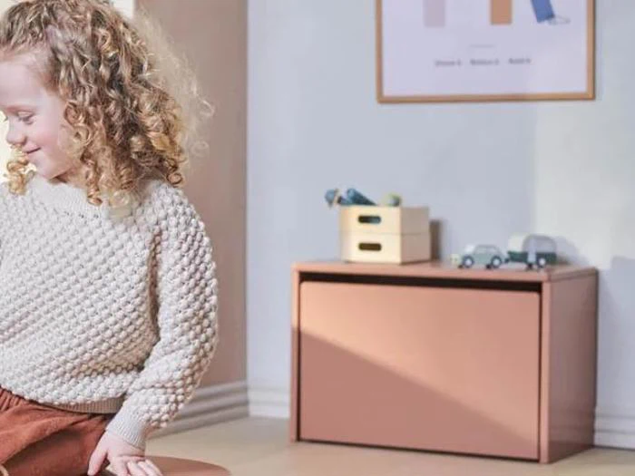 Et lille barn med krøllet hår og en struktureret sweater sidder på en skammel i et svagt oplyst rum med pastelfarvede møbler og legetøjsbiler på et skab i baggrunden.