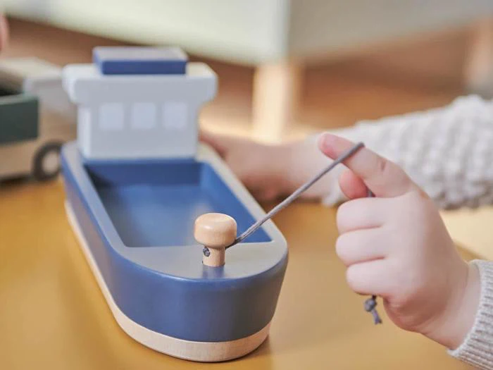 En barnehånd trækker i en snor, der er fastgjort til en blå legetøjsbåd af træ på et lysebrunt bord.