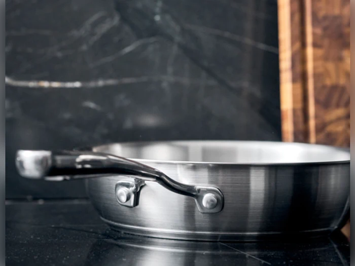 En stegepande af rustfrit stål med håndtag står på en bordplade af sort marmor med et skærebræt af træ i baggrunden.